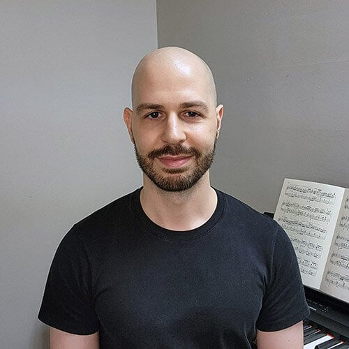 David Miele, respected piano teacher and owner, sits indoors in a black T-shirt. Beside him is a music stand holding open sheet music against the grey wall.